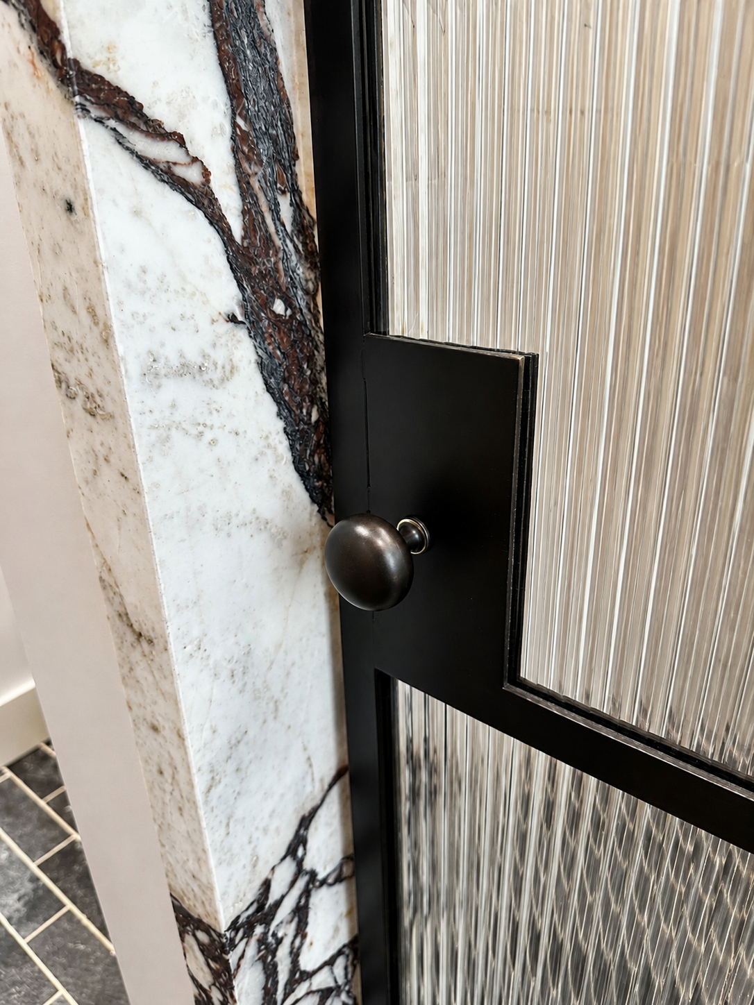 Close detail of black metal ribbed glass shower door handle and marble surround