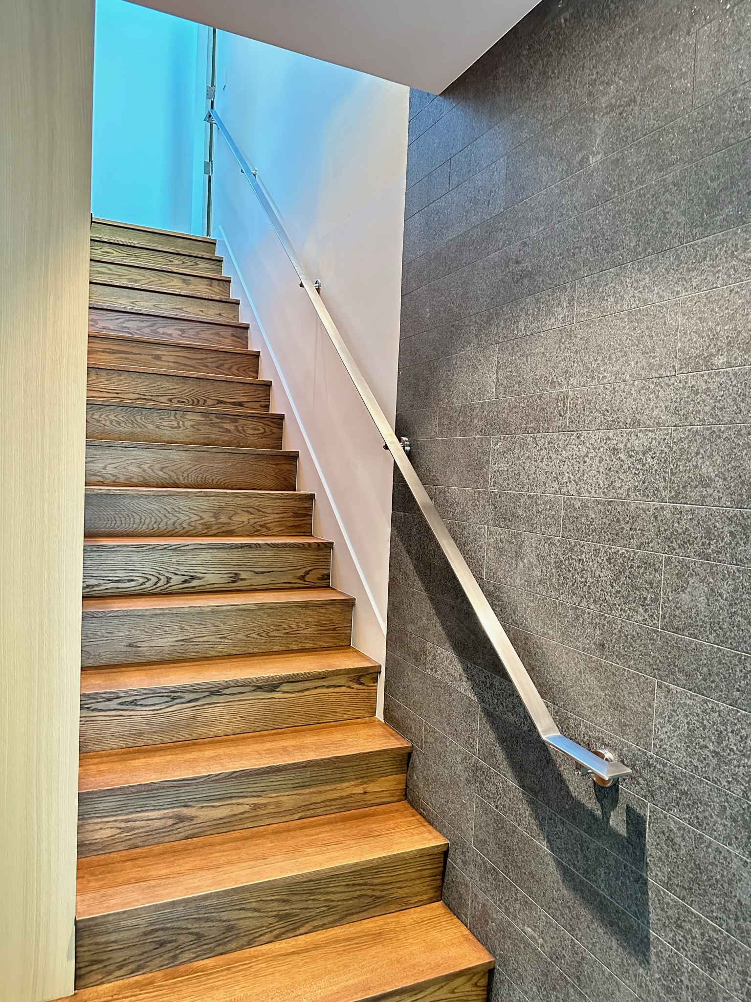 Stainless steel stair handrail beside wood steps and gray tile wall