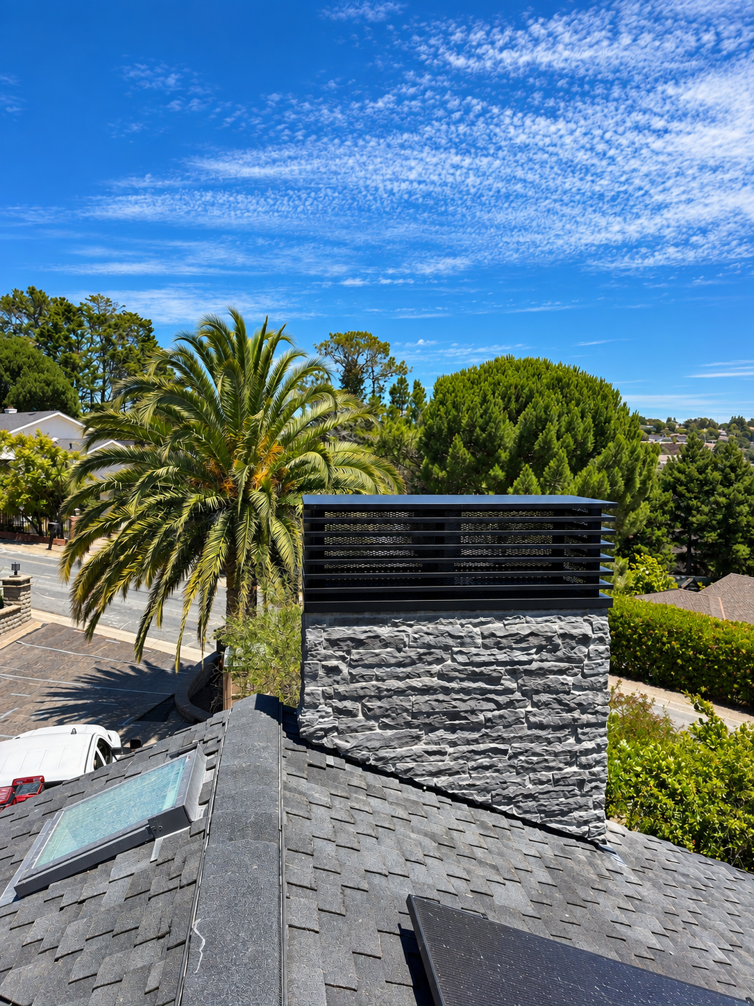 Black metal chimney cap on a stone chimney with hillside trees and blue sky