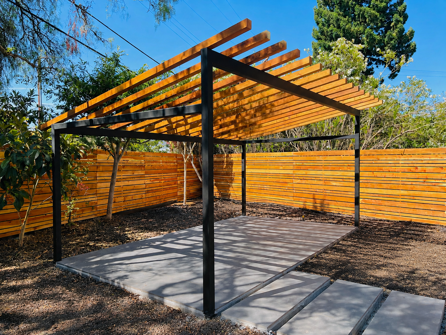 Wide view of black metal pergola with wood slat roof and matching wood fence